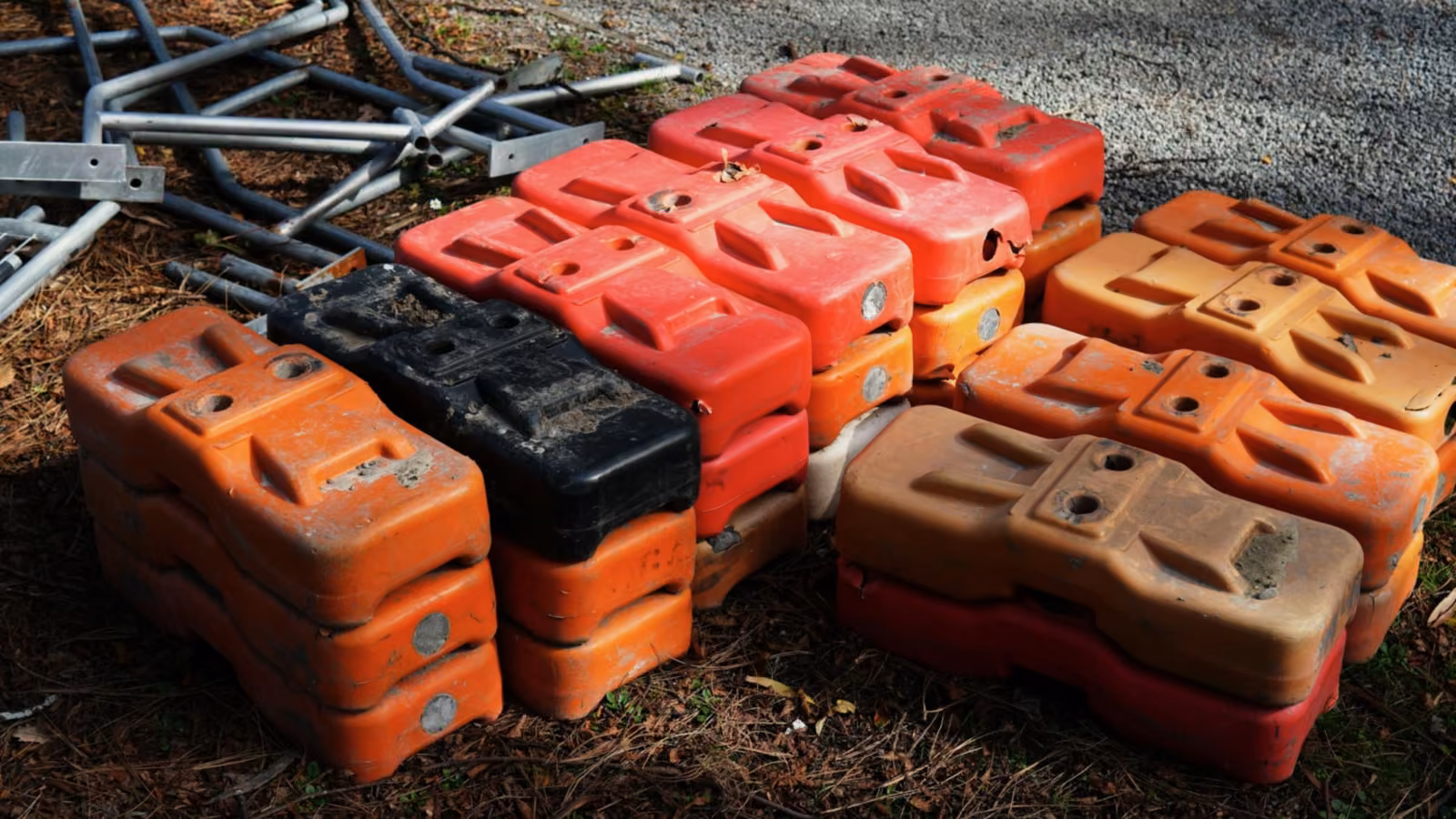 Gestapelte Bauzaunfüße aus Kunststoff in Orange, Rot und Schwarz, dahinter abgelegte verzinkte Stahlrohrrahmen.