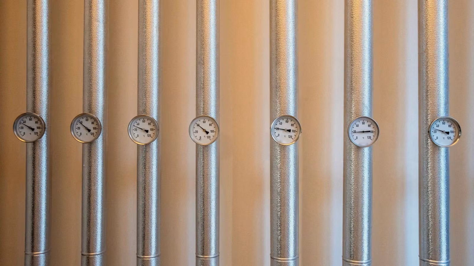 Senkrechte, mit Aluminium ummantelte Heizungsleitungen in einer Technikzentrale, an jeder Leitung ein Zeigerthermometer.