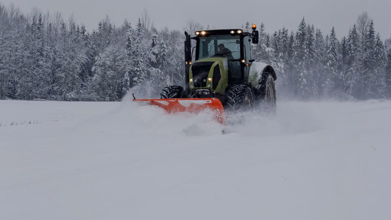 Traktor mit rotem Frontschild räumt Schnee auf einem weiten Feld vor einem verschneiten Waldrand.